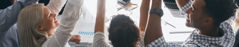 Portrait from back of glad students sitting together at the table and raising hands. Indoor photo of team of freelance specialists having fun after hard work in office.